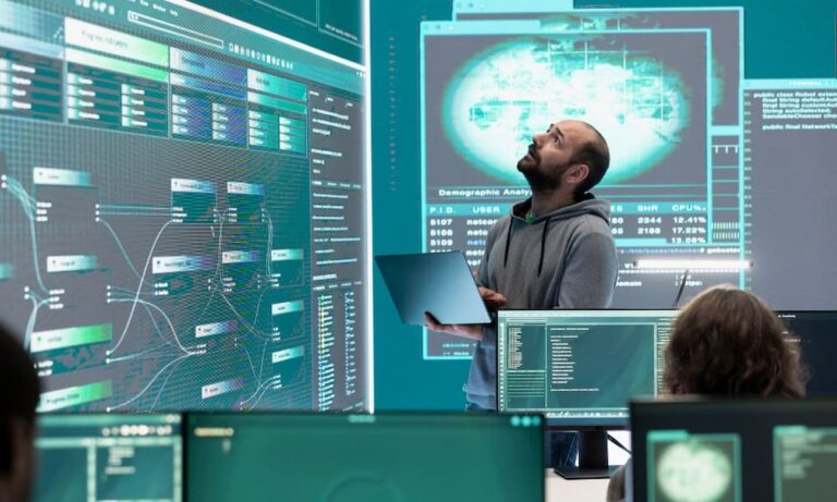 An IT hacker specialist reviews cybersecurity and recovery on a large screen while holding a laptop.