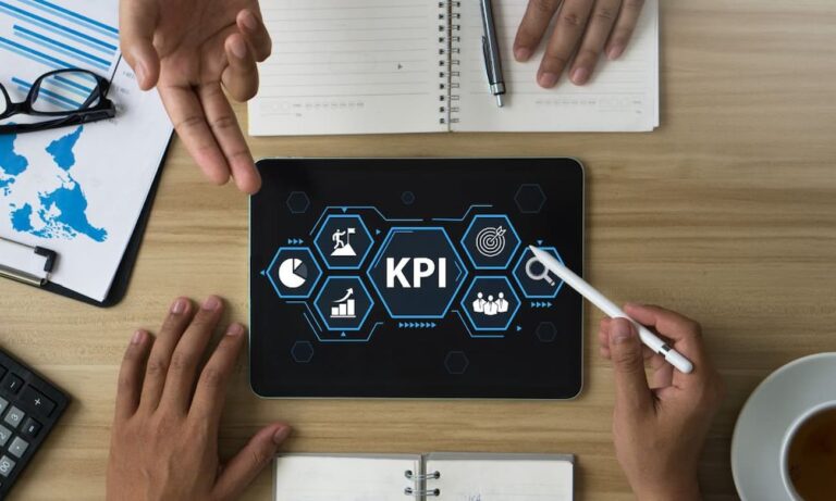 Two business people sit at a table with open notebooks. They are looking at a digital tablet displaying key performance indicators.