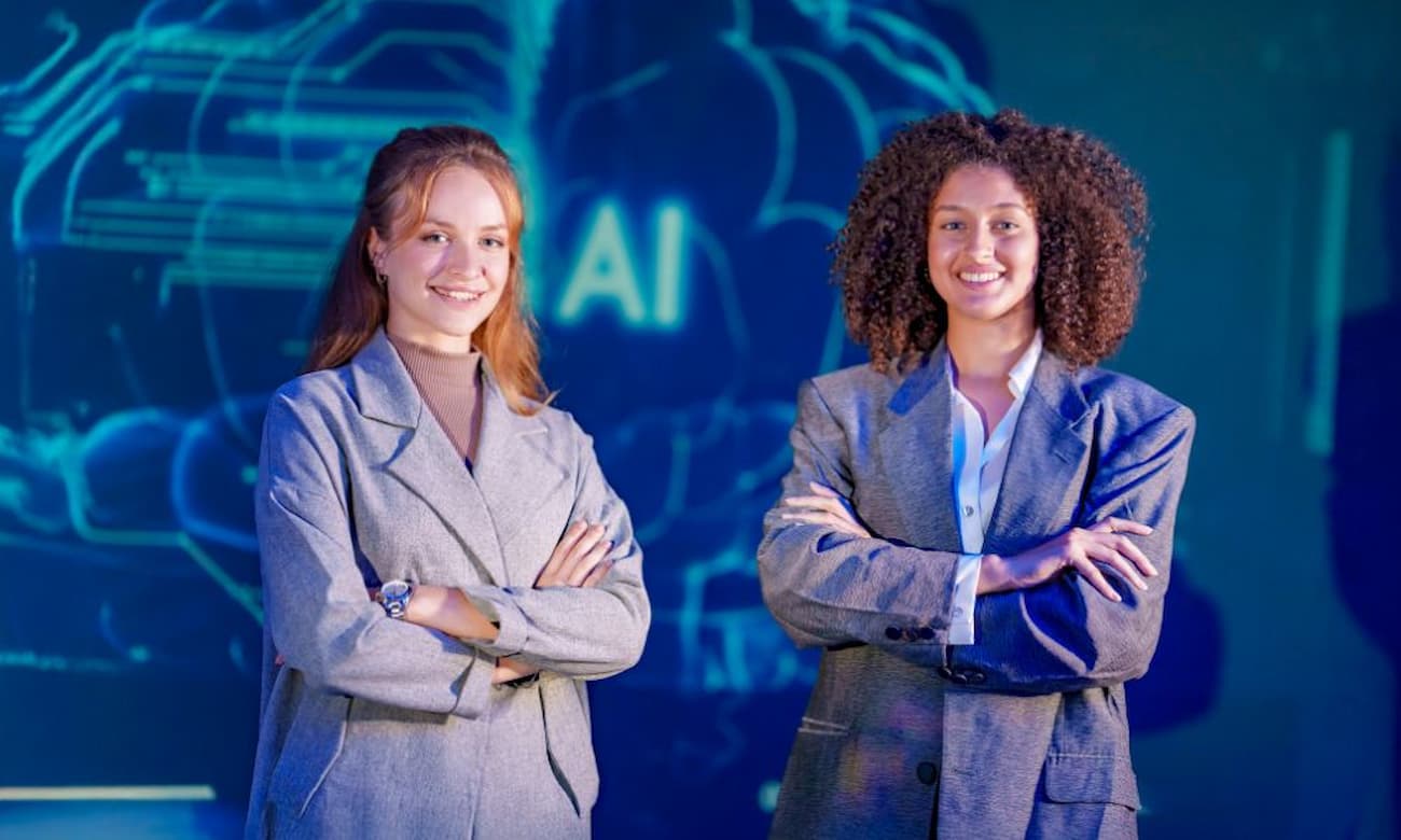 Two young women stand confidently in a modern workplace, arms crossed, against a vibrant blue backdrop featuring an 'AI' design.