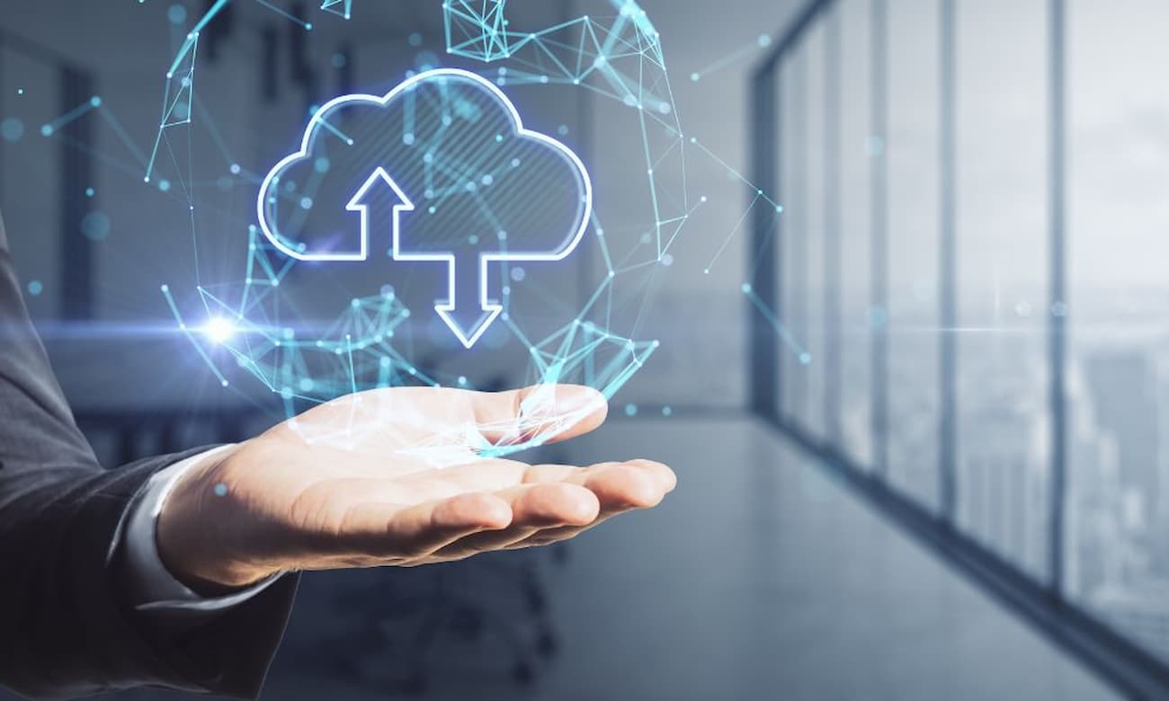 A hand in a suit jacket presents a glowing cloud computing graphic against a blurred office interior. The room is lit with bright, diffused light, and the futuristic rendering features geometric design elements and blue hues.