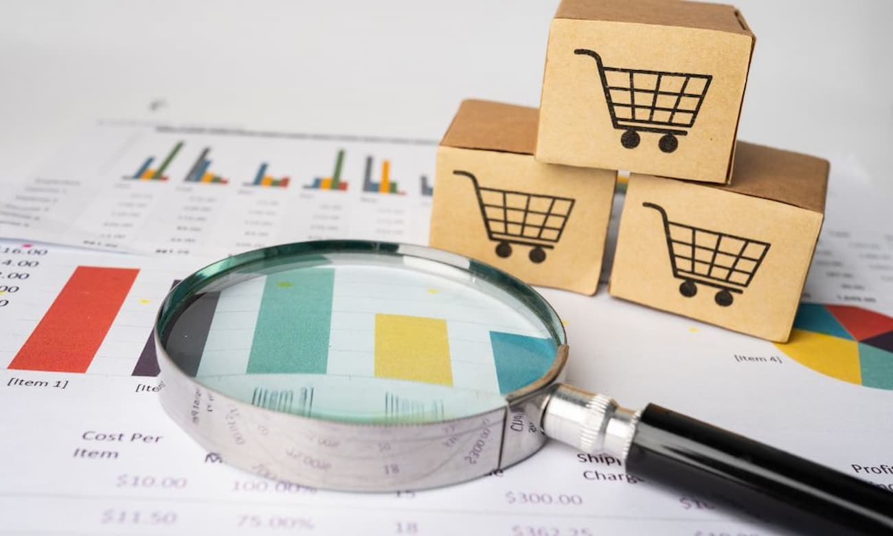 An eye-level shot depicts a magnifying glass, paper with colorful graphs and charts, and three small brown cardboard boxes with black shopping cart images stacked in a pyramid. The scene evokes themes of data analysis, retail trends, and business growth in the marketplace.