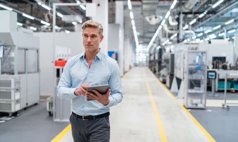 A man with short blond hair and wearing a blue collared shirt and gray pants is walking down a long corridor while holding a tablet in a factory. The factory appears clean and well-maintained, indicating a sophisticated, modern approach to industry.