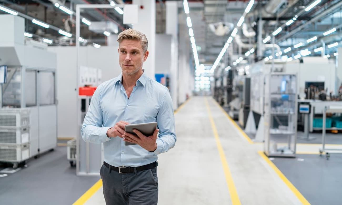 A man with short blond hair and wearing a blue collared shirt and gray pants is walking down a long corridor while holding a tablet in a factory. The factory appears clean and well-maintained, indicating a sophisticated, modern approach to industry.