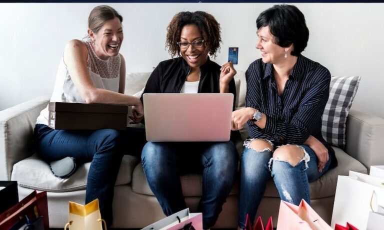 Three smiling women sit on a sofa together in the daytime. One woman holds a credit card while looking at a pink laptop on her lap. Another woman holds a shoebox, and shopping bags are arranged on the floor.