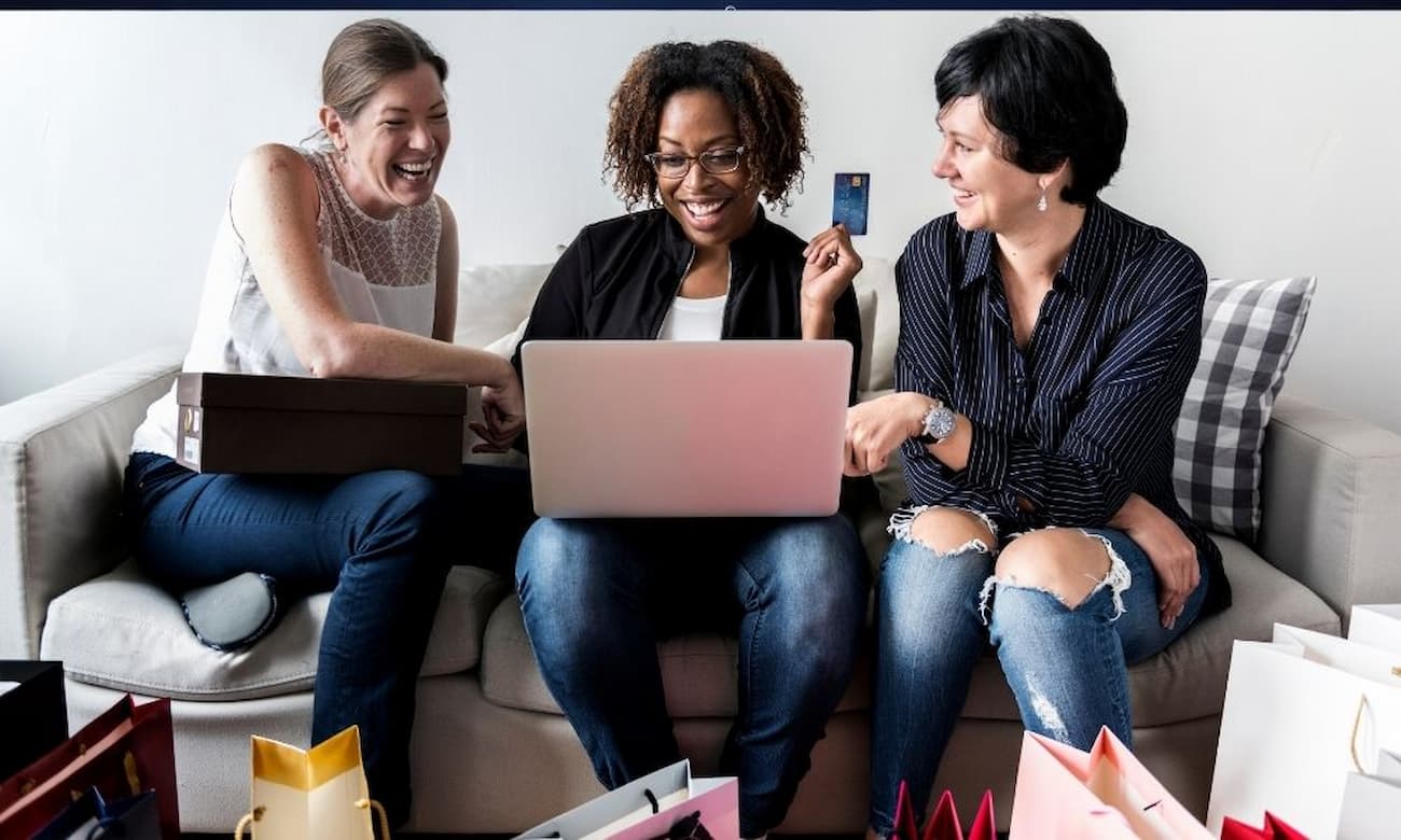 Three smiling women sit on a sofa together in the daytime. One woman holds a credit card while looking at a pink laptop on her lap. Another woman holds a shoebox, and shopping bags are arranged on the floor.