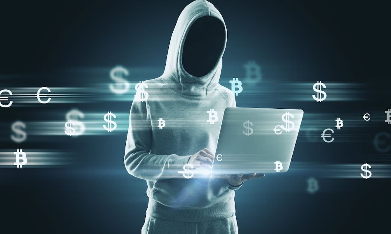 A hooded person uses a laptop surrounded by digital currency symbols.